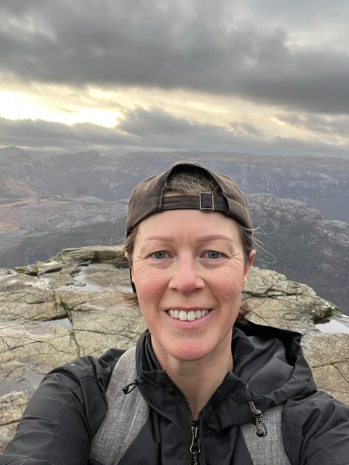 A white woman wearing a backwards hat and backpack smiles atop a rocky landscape.